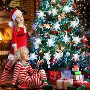 Two children in festive pajamas sitting on the floor in front of a decorated Christmas tree with presents underneath.