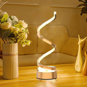 Spiral table lamp on a wooden surface with flowers and a vase in the background