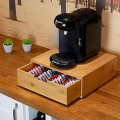 Black coffee machine on a wooden stand with a drawer containing coffee pods, set against a wooden wall.