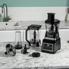 Black food processor with attachments on a marble countertop in a kitchen.