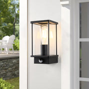Black outdoor wall light fixture on a white wall with greenery in the background
