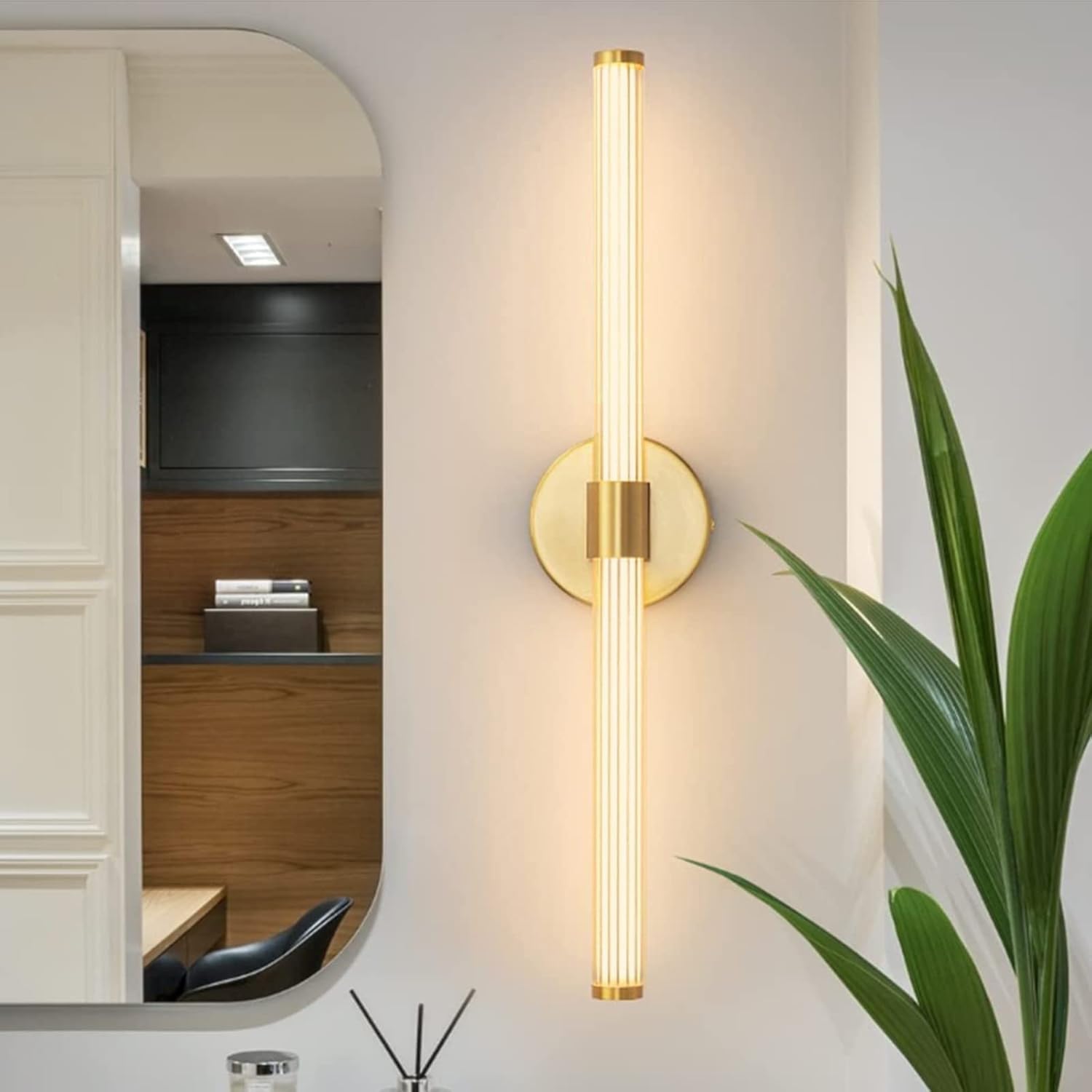 Gold wall sconce on a white wall with a mirror and plant in the background