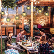 Group of people sitting around a table in an outdoor setting with string lights and plants.