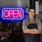 Man wearing an apron standing in front of a neon 'OPEN' sign inside a restaurant.