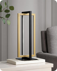 Modern table lamp with black and gold design on a white surface, gray wall, and gray sofa in the background.