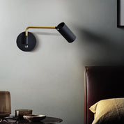 Modern wall lamp with black and gold design on a gray wall next to a bed.