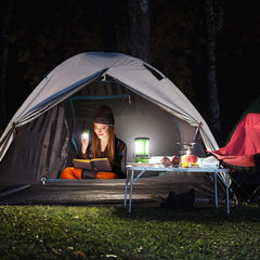 Person reading a book inside a tent with a lantern, surrounded by camping gear.