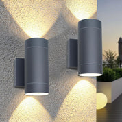 Two cylindrical outdoor wall lights on a textured stone wall with a blurred garden background.