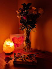 Vase with flowers, salt lamp, and heart-shaped object on a wooden surface with warm lighting.