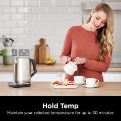 Woman pouring tea into a cup in a kitchen with a kettle and food on the counter.