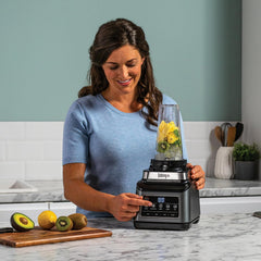 Woman using a Ninja blender with fruits on a kitchen counter