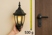 Hand holding a black outdoor wall light fixture next to a door with a measuring tape showing weight.